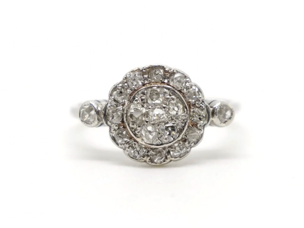 An Art Deco platinum cluster ring set with old cut diamonds arranged in a circular floral pattern with accent diamonds on the shoulders.
