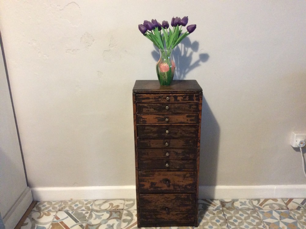 A tall and narrow dark brown wooden chest featuring nine graduated drawers with small brass knobs and a distressed finish. View II