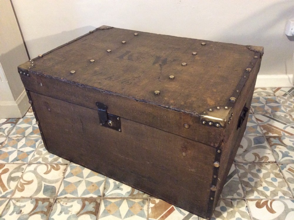 A rectangular Victorian dark brown canvas-covered trunk featuring brass corner caps, brass studs, a central lock, and stencilled black initials on the flat lid. View X