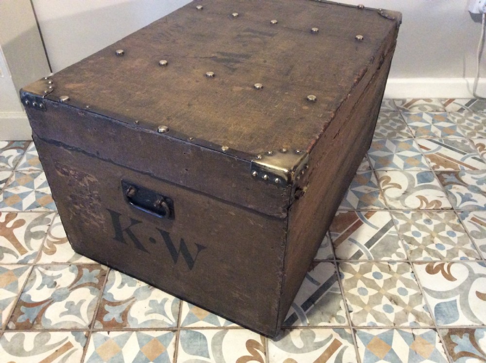 A rectangular Victorian dark brown canvas-covered trunk featuring brass corner caps, brass studs, a central lock, and stencilled black initials on the flat lid. View XX