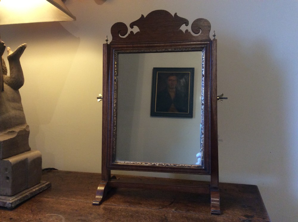 A Georgian dark mahogany skeleton toilet mirror featuring a scroll-carved cresting, rectangular pivoting mirror, side brass fixings, and trestle feet.
