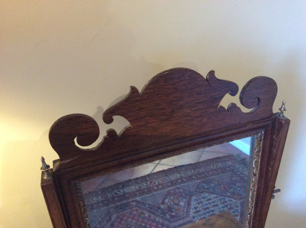 A Georgian dark mahogany skeleton toilet mirror featuring a scroll-carved cresting, rectangular pivoting mirror, side brass fixings, and trestle feet. View III