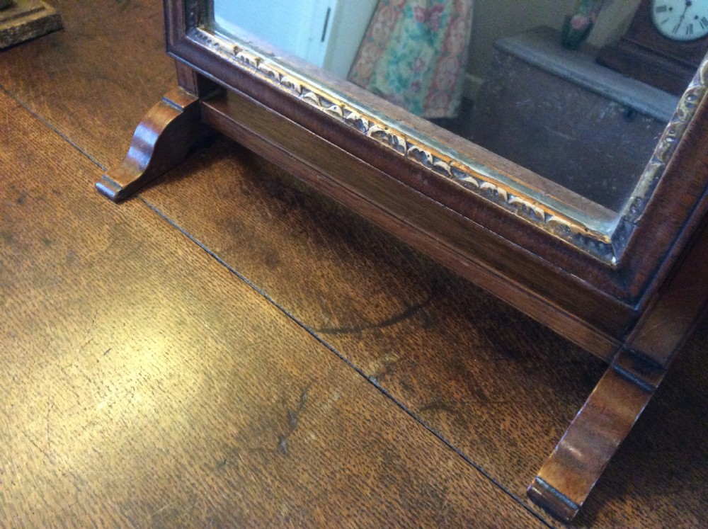 A Georgian dark mahogany skeleton toilet mirror featuring a scroll-carved cresting, rectangular pivoting mirror, side brass fixings, and trestle feet. View VI