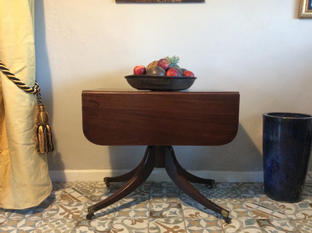 A Georgian mahogany drop-leaf hall table with two hinged side flaps, supported by a central pedestal and four curved splayed legs with brass castors.