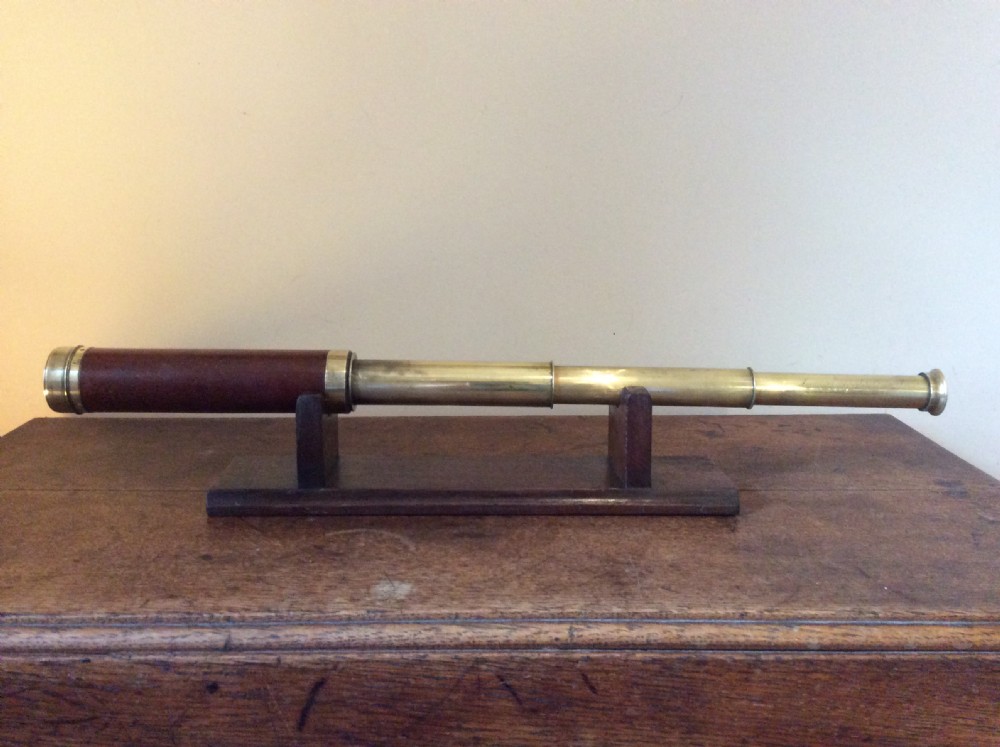 A Regency era brass multi-draw pocket telescope with a brown main barrel resting horizontally on a dark timber display stand.