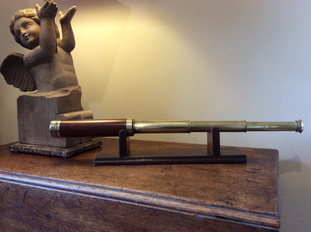 A Regency era brass multi-draw pocket telescope with a brown main barrel resting horizontally on a dark timber display stand. View II