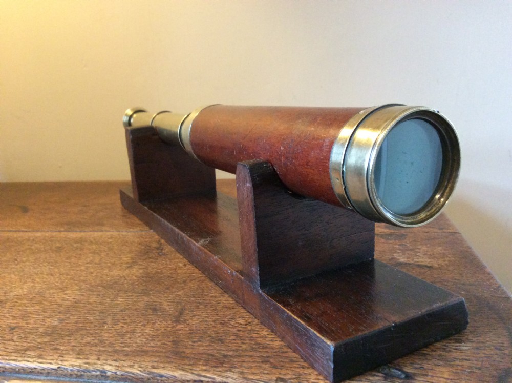 A Regency era brass multi-draw pocket telescope with a brown main barrel resting horizontally on a dark timber display stand. View VI