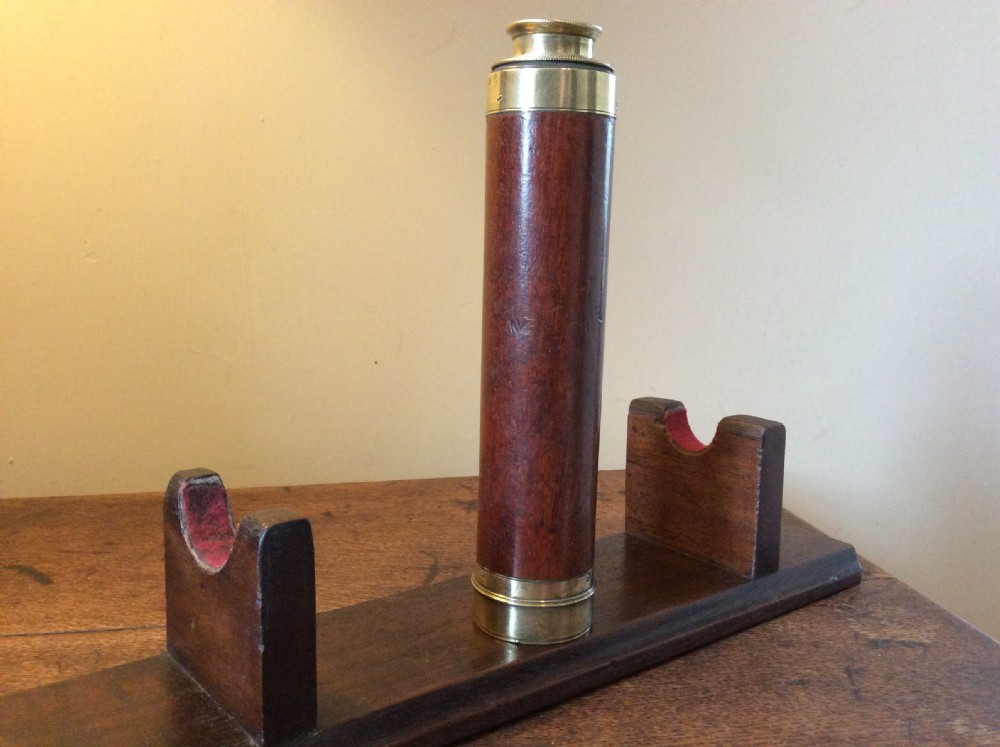 A Regency era brass multi-draw pocket telescope with a brown main barrel resting horizontally on a dark timber display stand. View VII