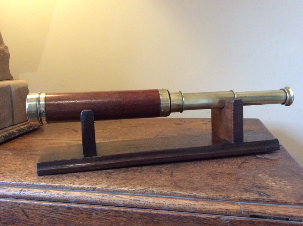 A Regency era brass multi-draw pocket telescope with a brown main barrel resting horizontally on a dark timber display stand. View VIII