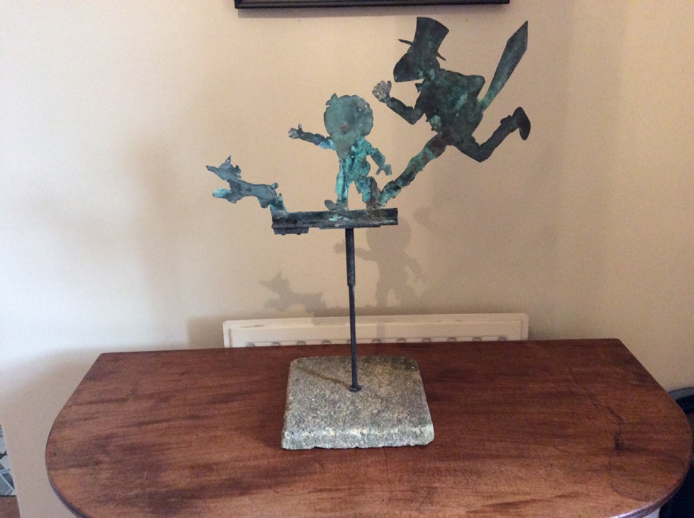 An early 20th-century copper weather vane featuring running figures with a natural verdigris patina, mounted on a stone block stand.