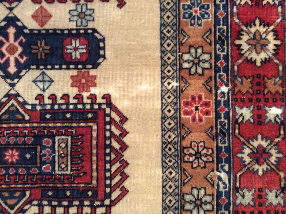 A small hand-woven antique Anatolian wool rug featuring geometric central medallions on a cream field surrounded by a patterned red and navy border. View V