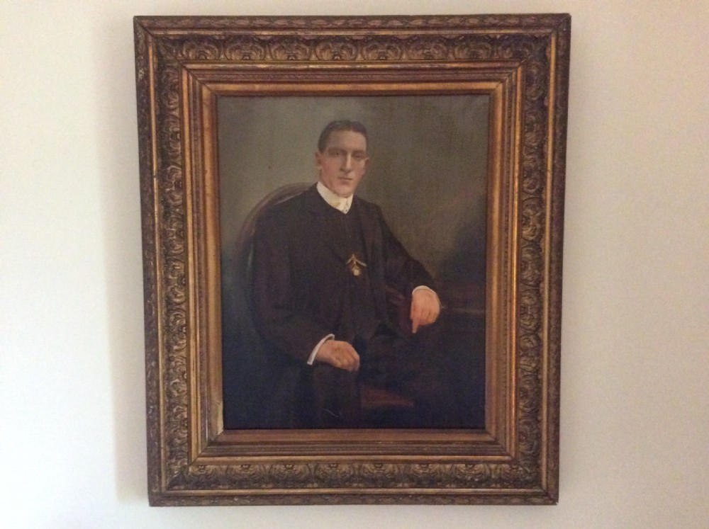 An antique oil painting depicting a seated gentleman in formal dark attire set within a heavily carved gold leaf frame.