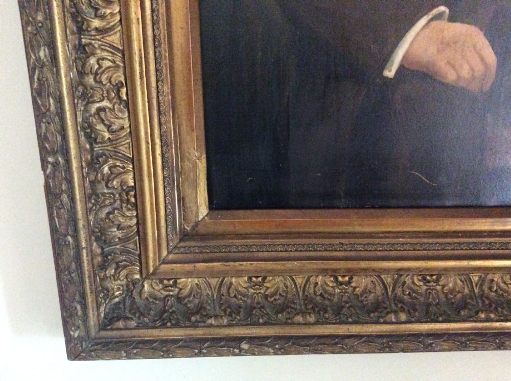 An antique oil painting depicting a seated gentleman in formal dark attire set within a heavily carved gold leaf frame. View IX