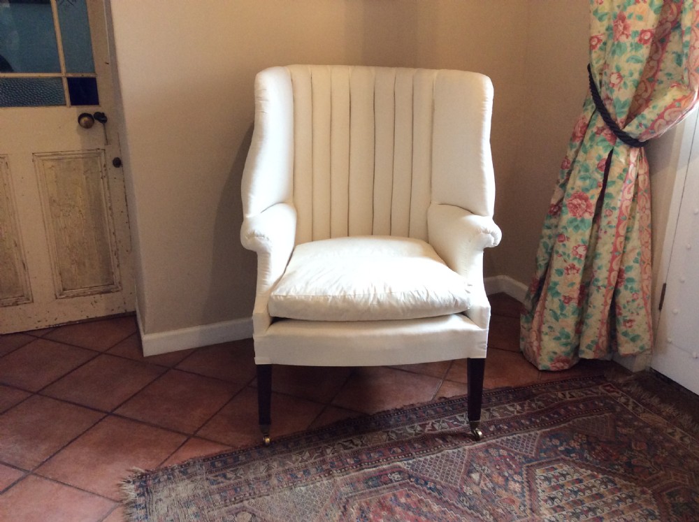 An antique George III high-backed barrel armchair upholstered in cream fabric with channel tufting, supported on dark mahogany legs with brass castors.