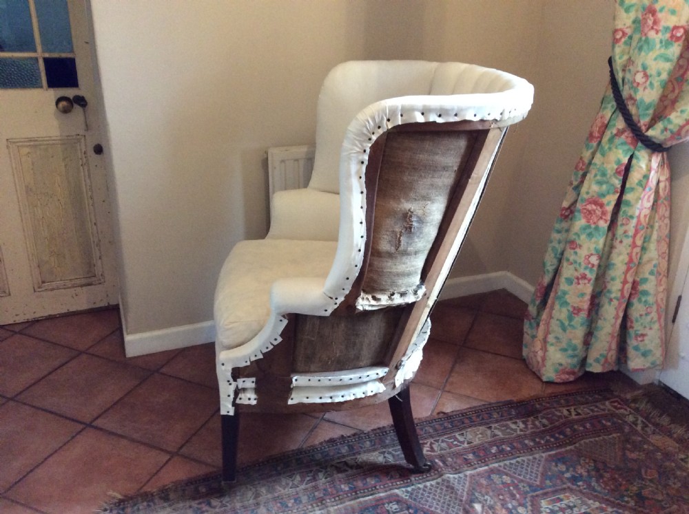 An antique George III high-backed barrel armchair upholstered in cream fabric with channel tufting, supported on dark mahogany legs with brass castors. View V