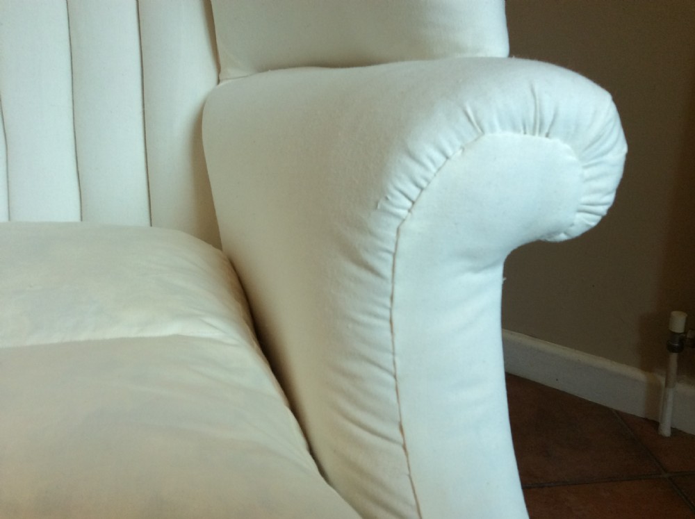 An antique George III high-backed barrel armchair upholstered in cream fabric with channel tufting, supported on dark mahogany legs with brass castors. View X