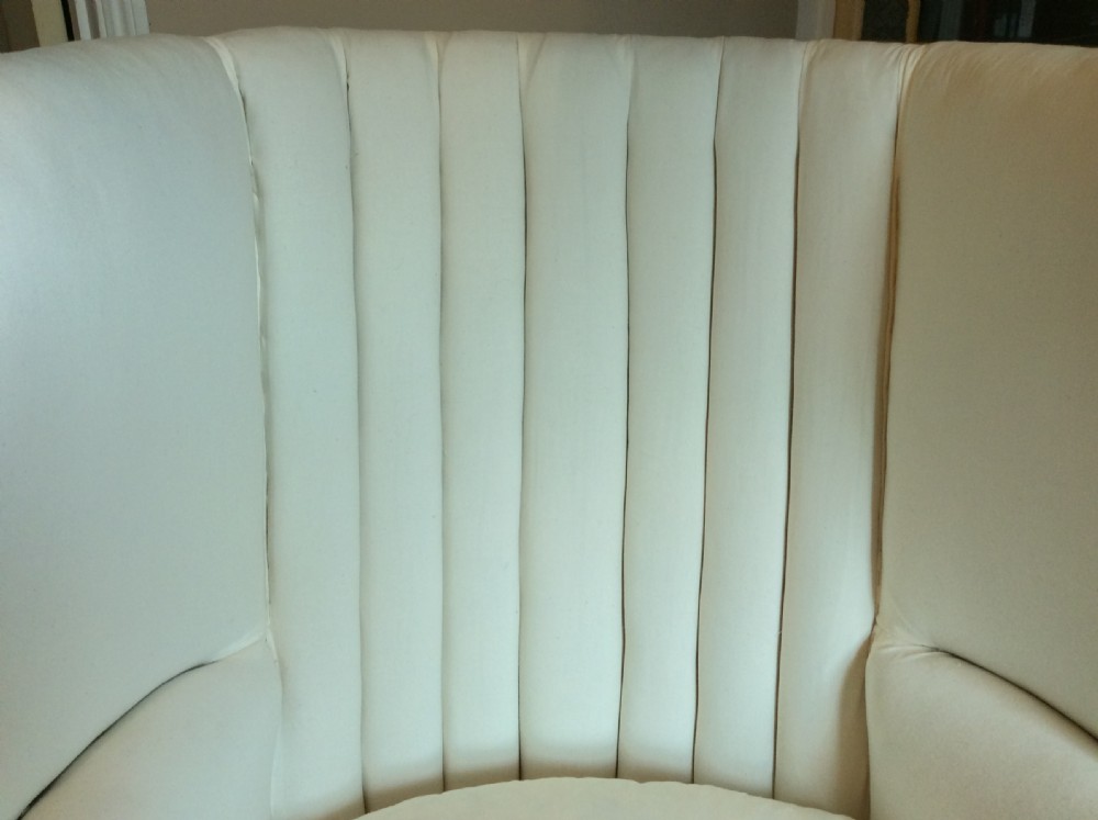 An antique George III high-backed barrel armchair upholstered in cream fabric with channel tufting, supported on dark mahogany legs with brass castors. View XI