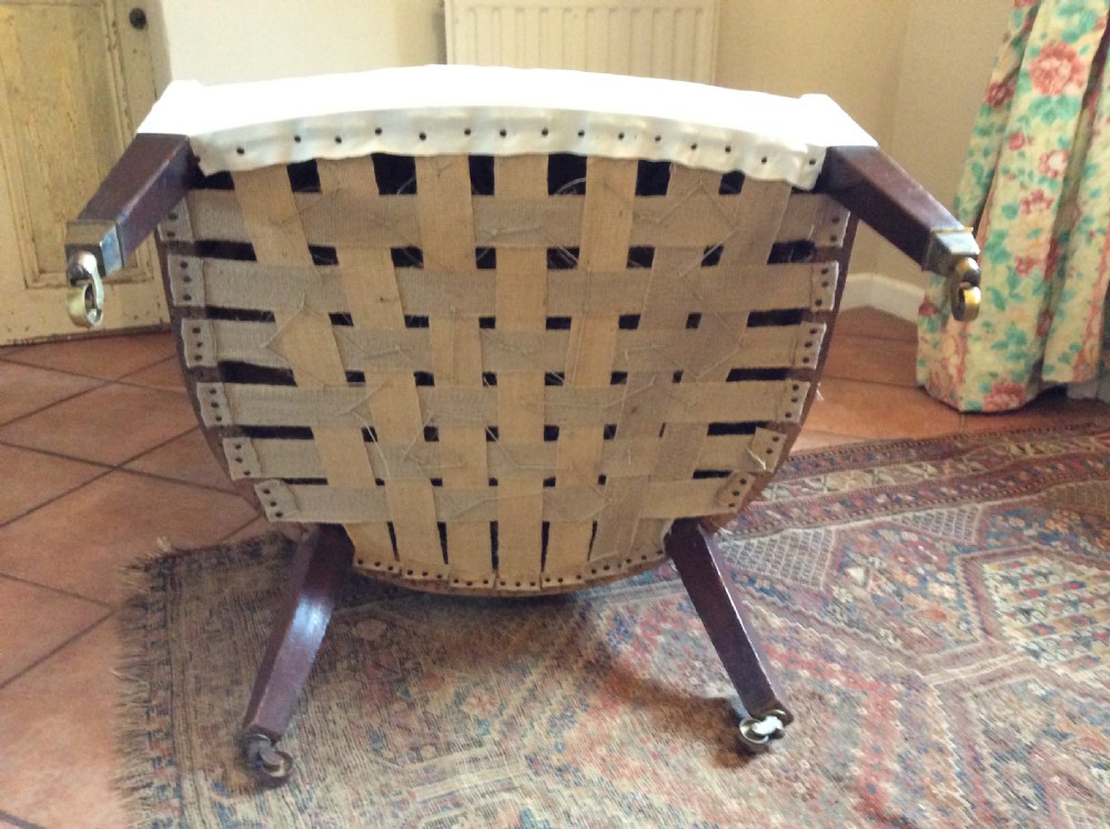 An antique George III high-backed barrel armchair upholstered in cream fabric with channel tufting, supported on dark mahogany legs with brass castors. View XII