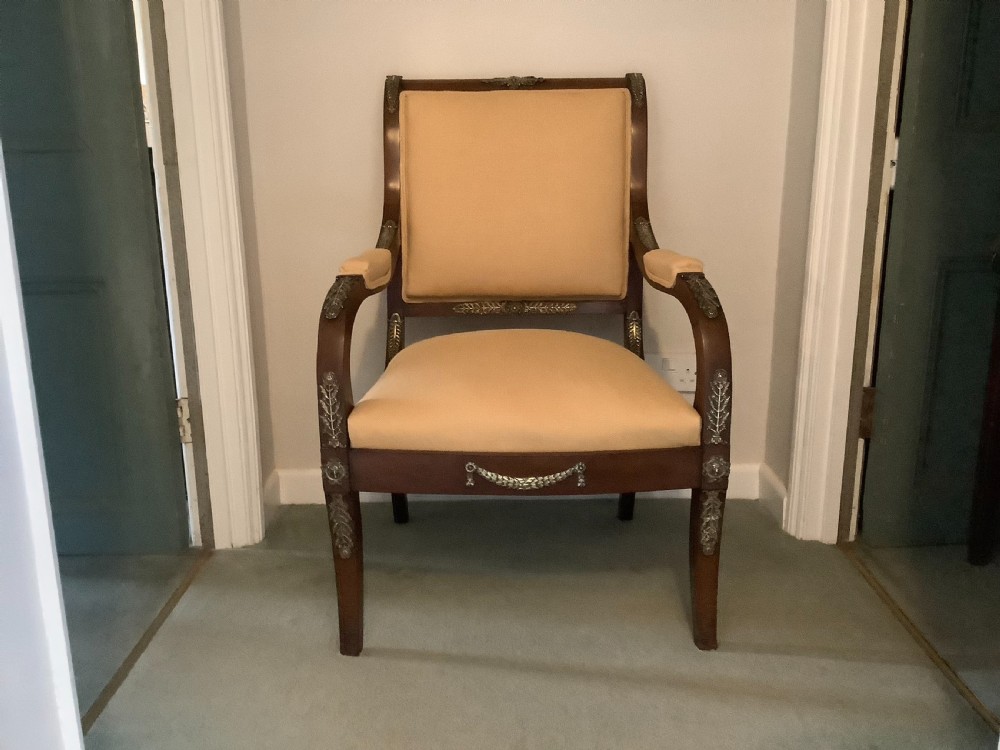 A 19th-century French Empire mahogany open armchair featuring light gold-coloured fabric upholstery and ornate brass leaf mounts.