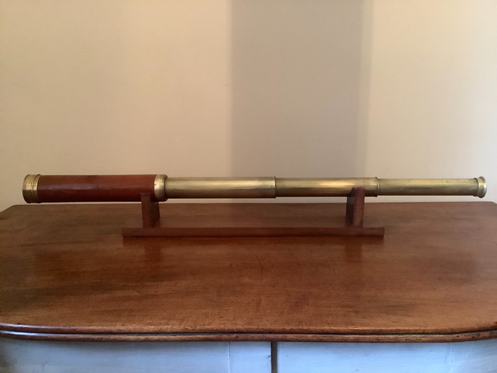 An antique brass multi-draw telescope with a leather-bound barrel rested horizontally on a fitted wooden display stand.