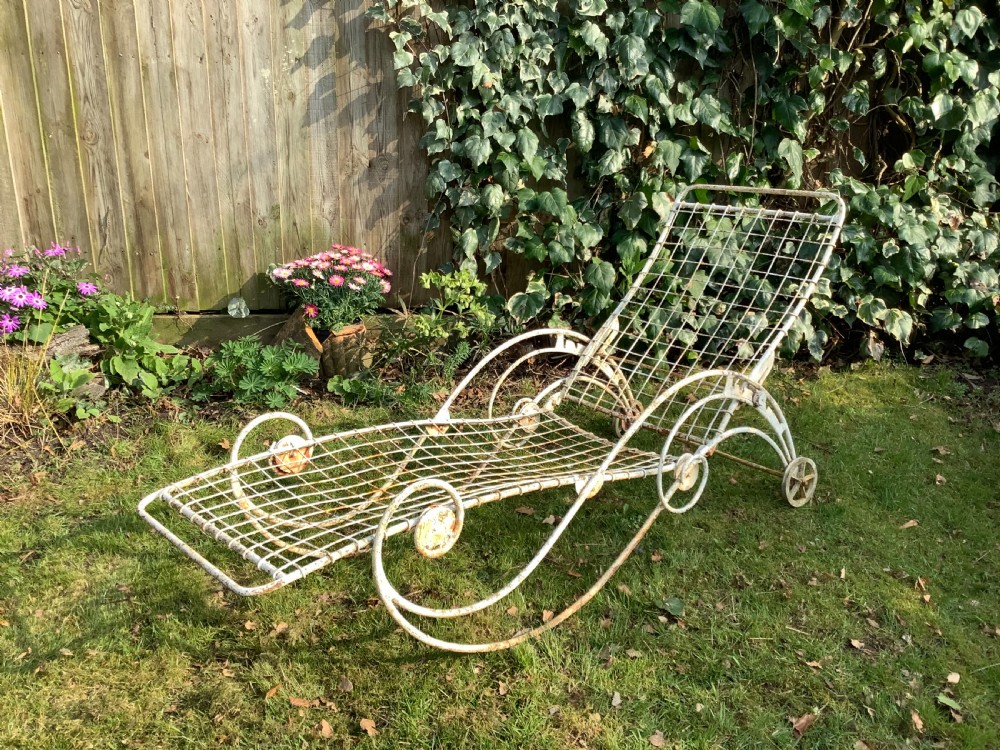 A weathered white wrought iron French garden lounger with a wire mesh reclining seat, curved scroll framework, and integrated wheels.