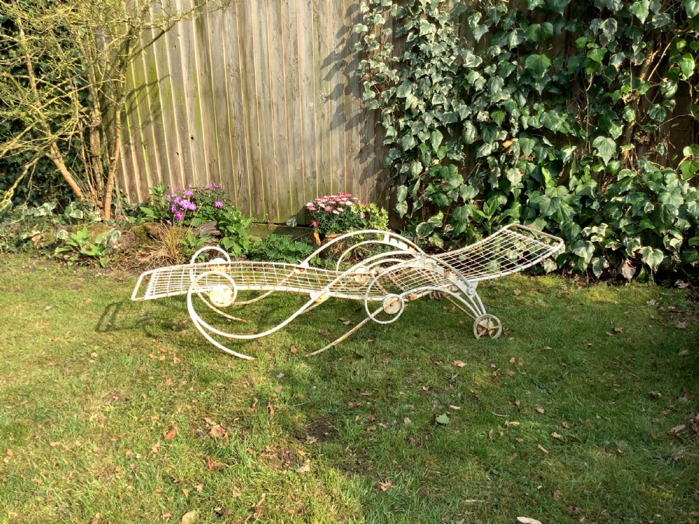 A weathered white wrought iron French garden lounger with a wire mesh reclining seat, curved scroll framework, and integrated wheels. View III