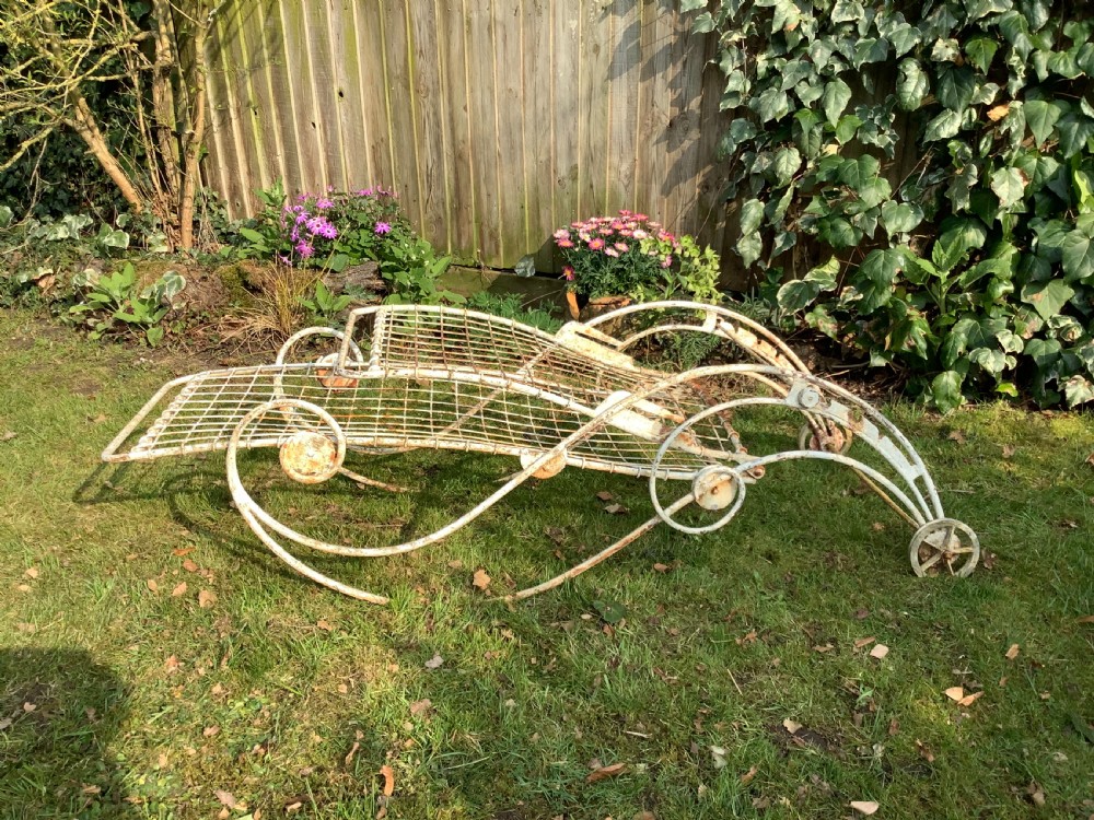 A weathered white wrought iron French garden lounger with a wire mesh reclining seat, curved scroll framework, and integrated wheels. View V