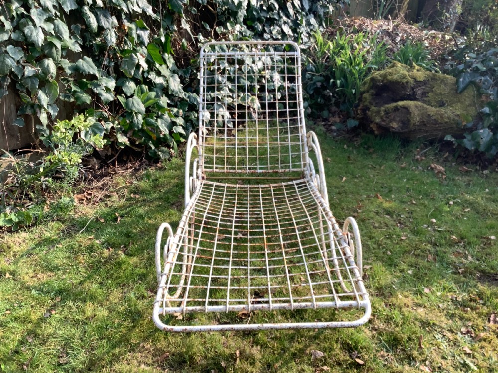 A weathered white wrought iron French garden lounger with a wire mesh reclining seat, curved scroll framework, and integrated wheels. View VI