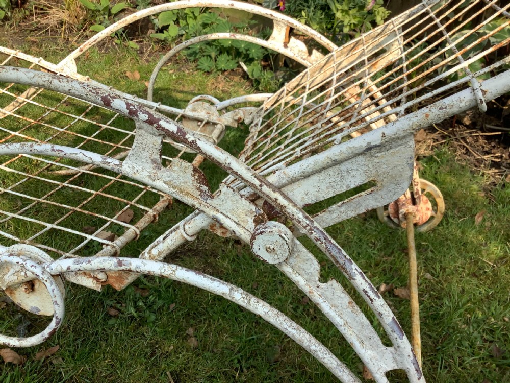 A weathered white wrought iron French garden lounger with a wire mesh reclining seat, curved scroll framework, and integrated wheels. View VII