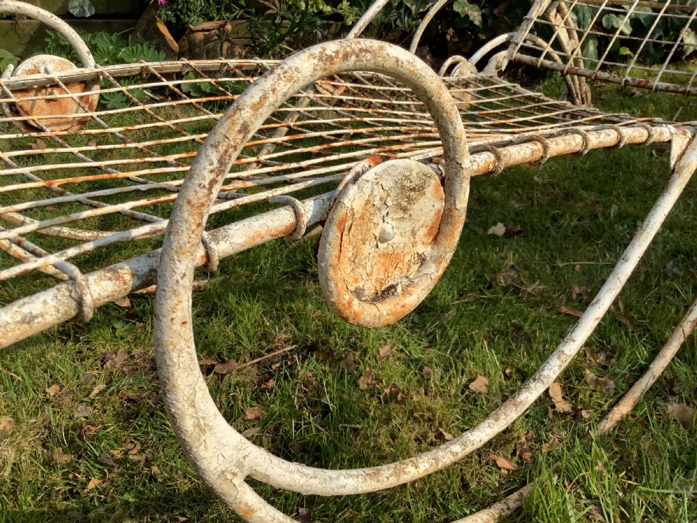 A weathered white wrought iron French garden lounger with a wire mesh reclining seat, curved scroll framework, and integrated wheels. View VIII