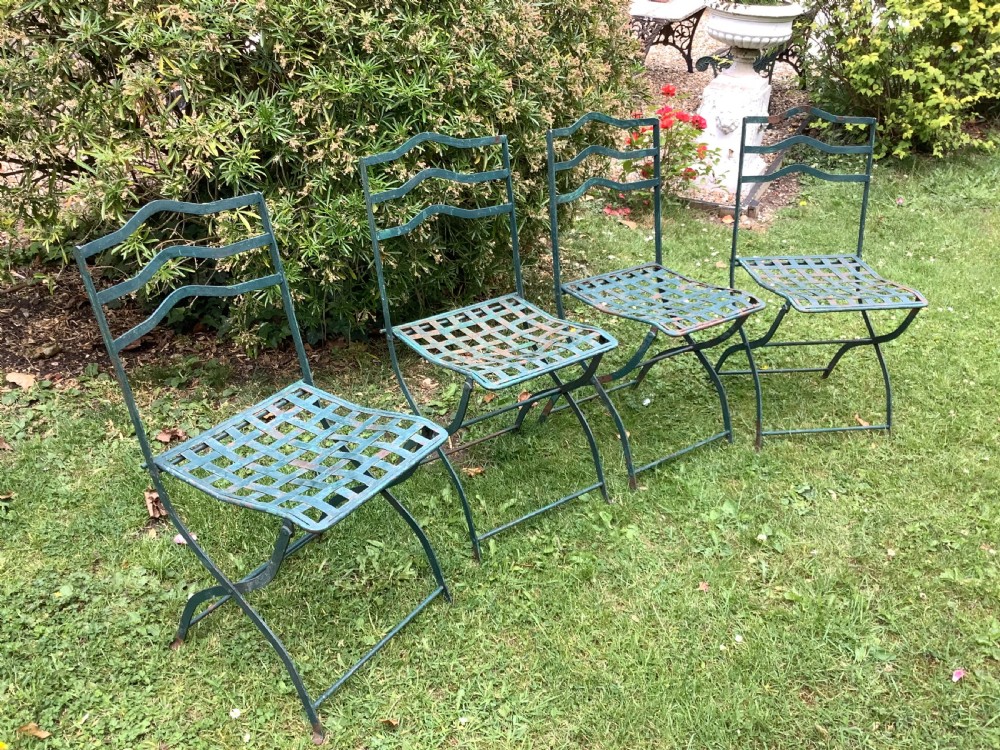 A set of four weathered green painted French folding iron garden chairs with lattice strap seats resting on grass.