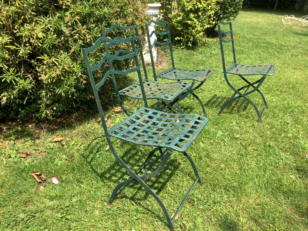 A set of four weathered green painted French folding iron garden chairs with lattice strap seats resting on grass. View III