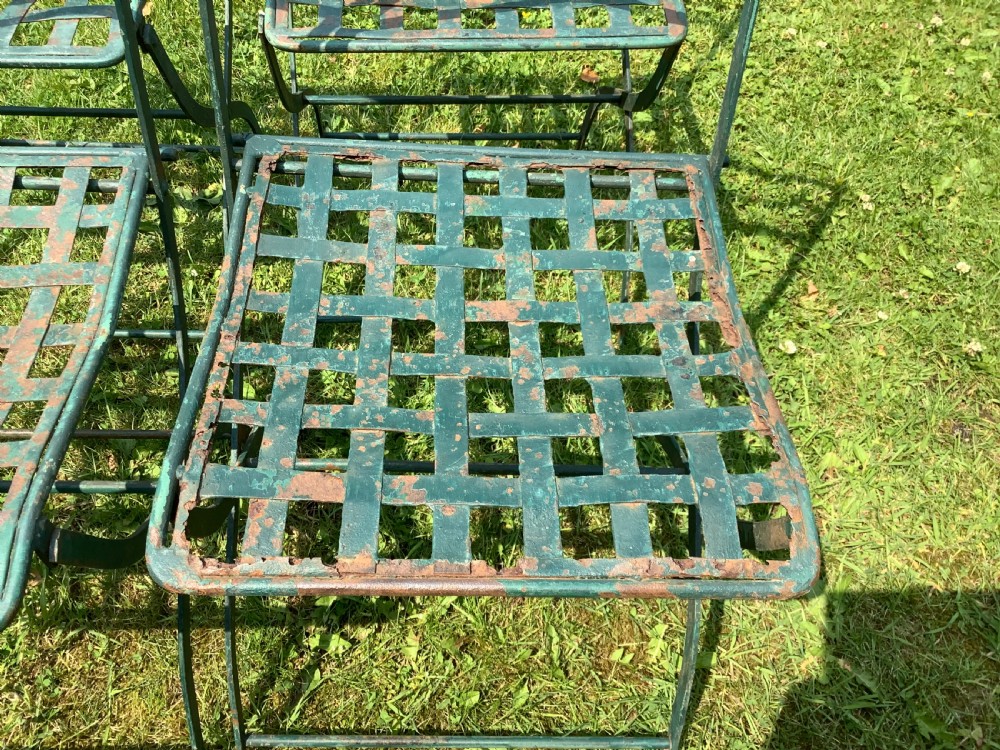 A set of four weathered green painted French folding iron garden chairs with lattice strap seats resting on grass. View IX