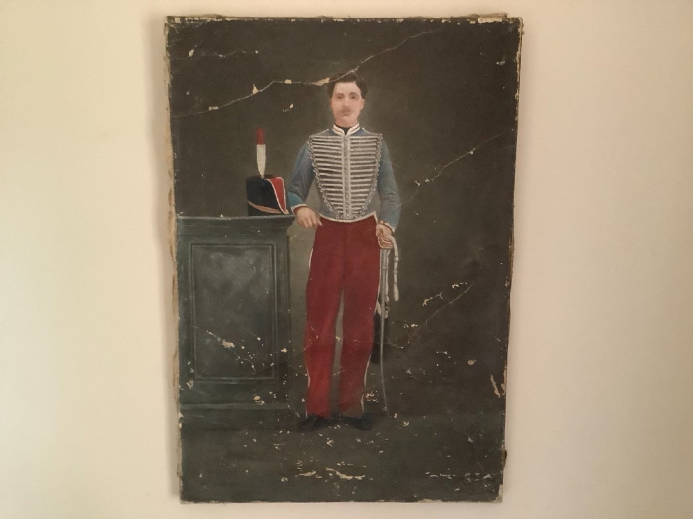 An antique full-length oil painting on canvas depicting a Hussar military officer standing in a braided blue uniform beside a pedestal.
