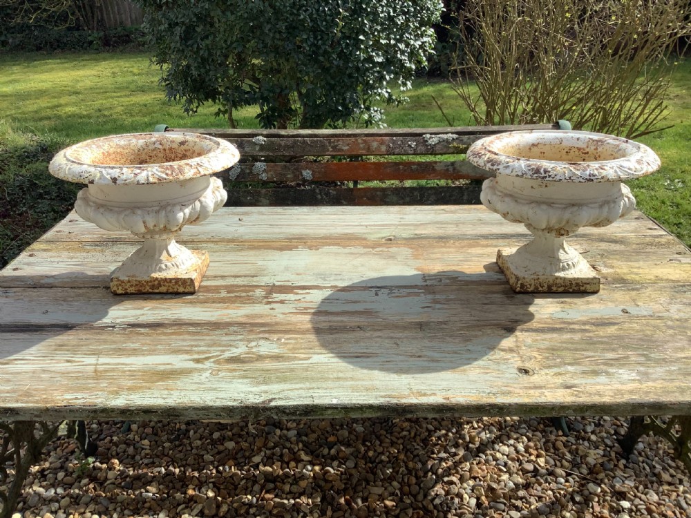 A pair of weathered off-white cast iron table urns featuring classical gadrooned decoration and square pedestal bases.