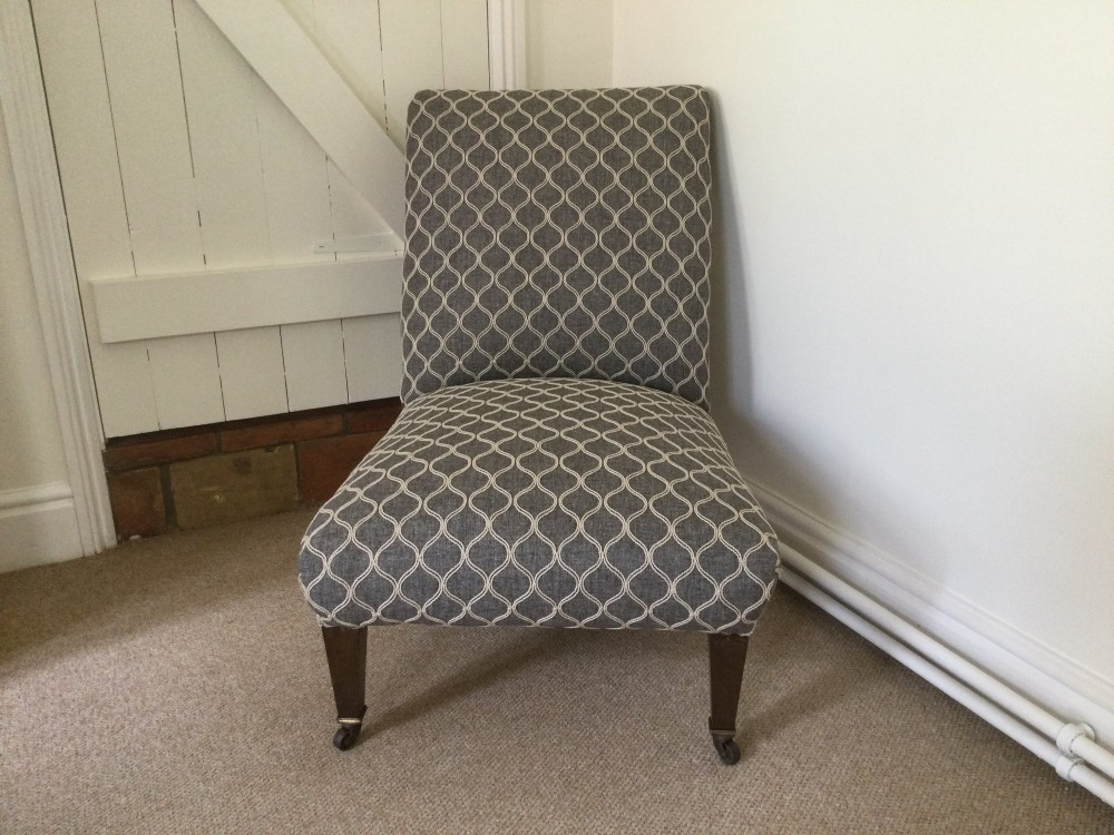 A 19th-century English low-seated slipper chair upholstered in grey patterned fabric with square tapered wooden legs on brass castors.