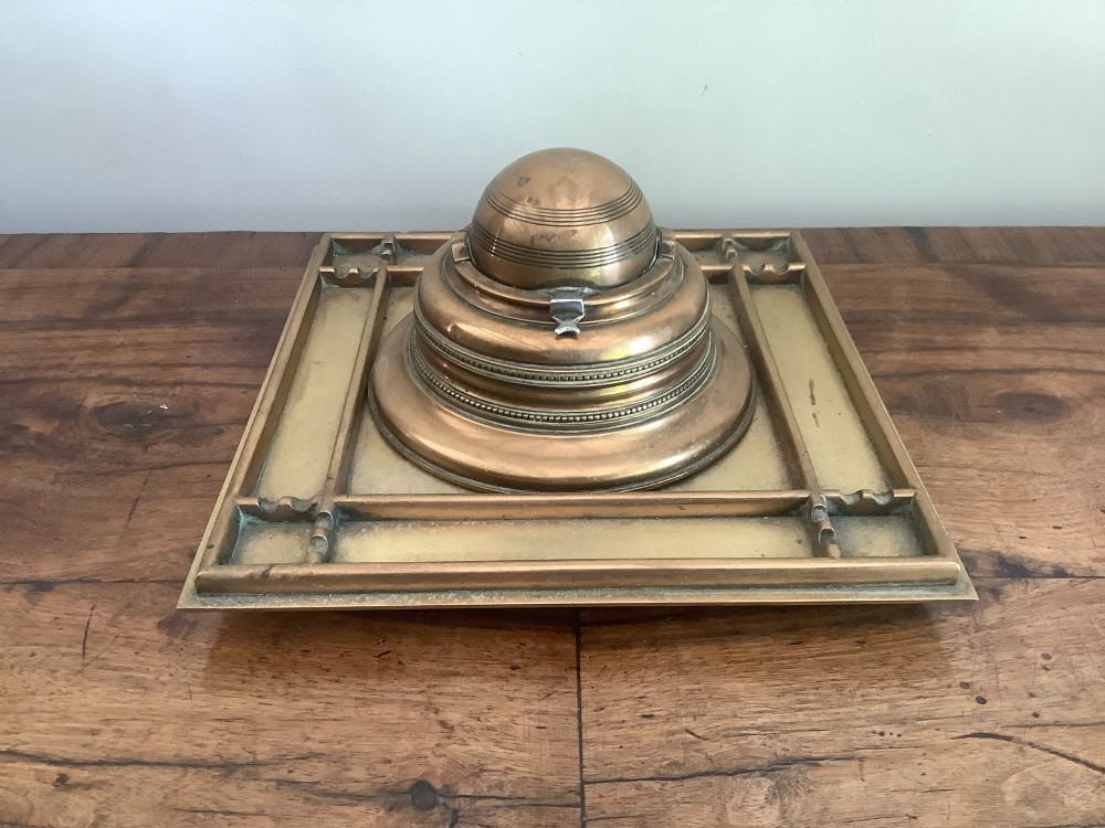 A square Victorian brass inkstand featuring a central domed hinged inkwell resting on a stepped base surrounded by pen channels.