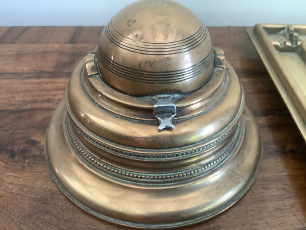 A square Victorian brass inkstand featuring a central domed hinged inkwell resting on a stepped base surrounded by pen channels. View V