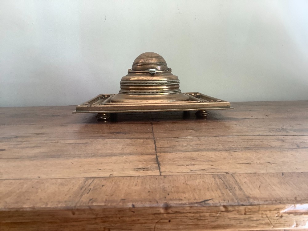 A square Victorian brass inkstand featuring a central domed hinged inkwell resting on a stepped base surrounded by pen channels. View VIII