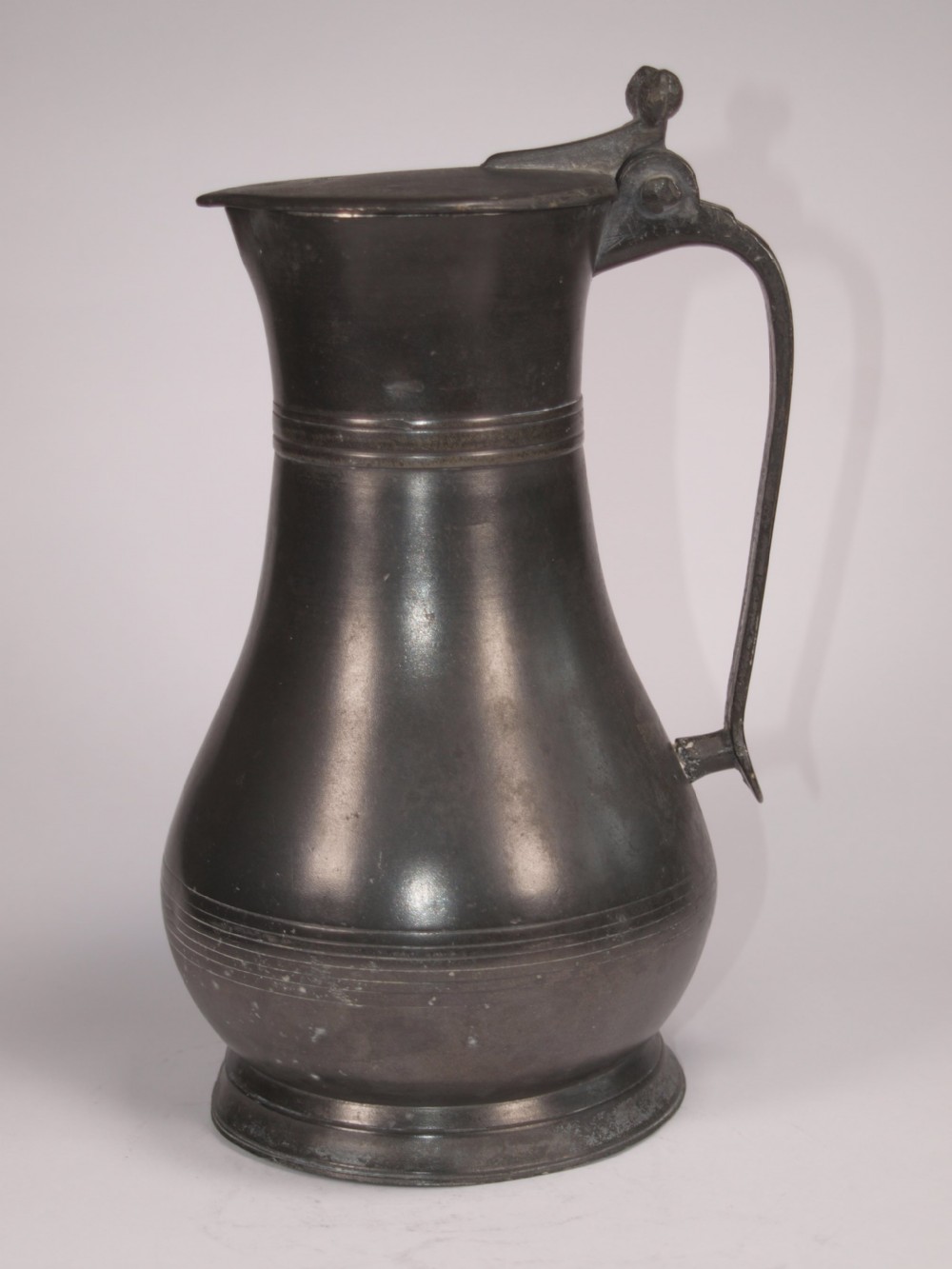 An antique dark grey pewter lidded flagon featuring a bulbous body, flared collar, hinged lid, and handle.
