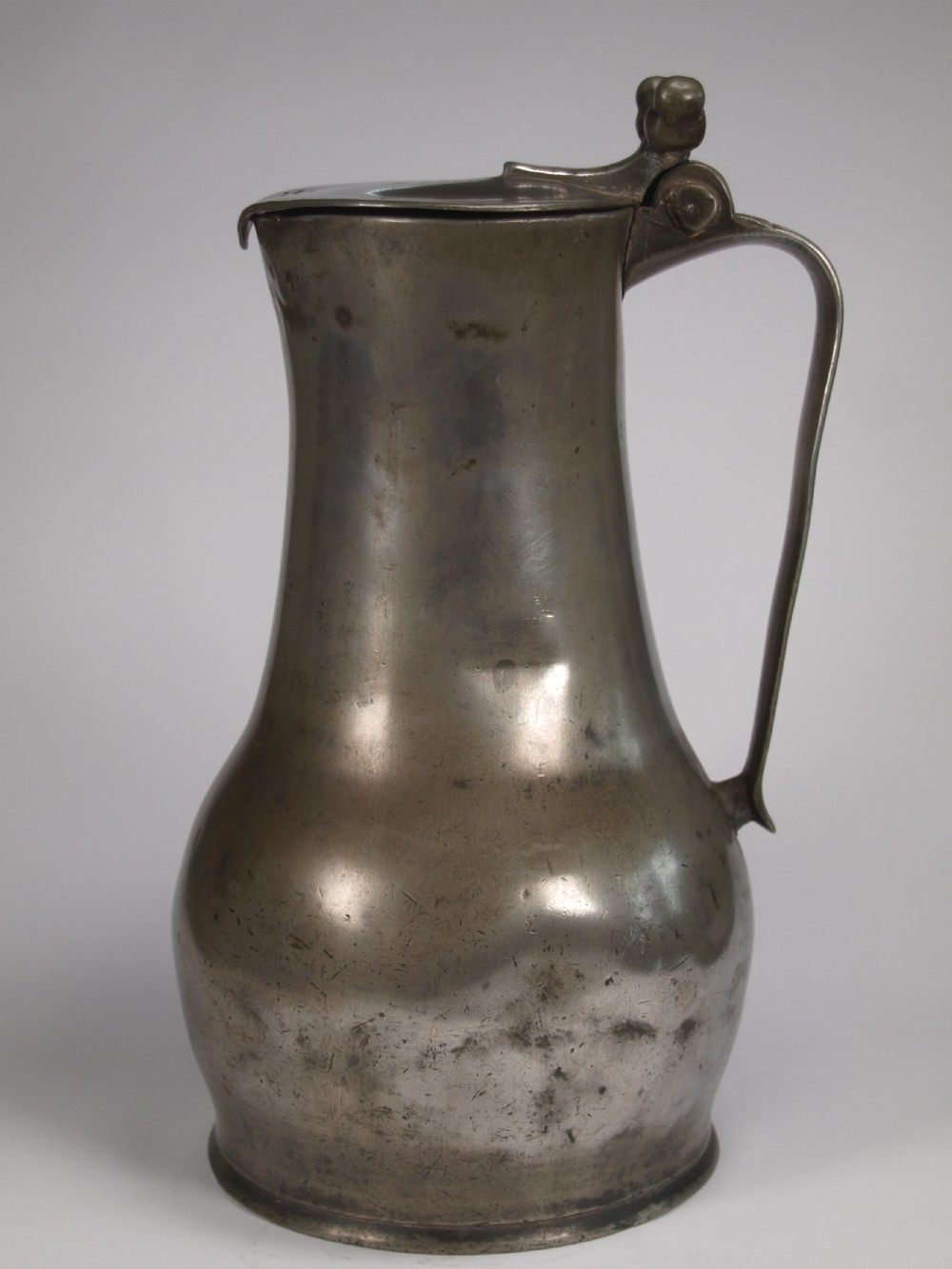 An antique pewter Jersey measure with a hinged lid, double acorn thumbpiece, curved handle, and bulbous body.