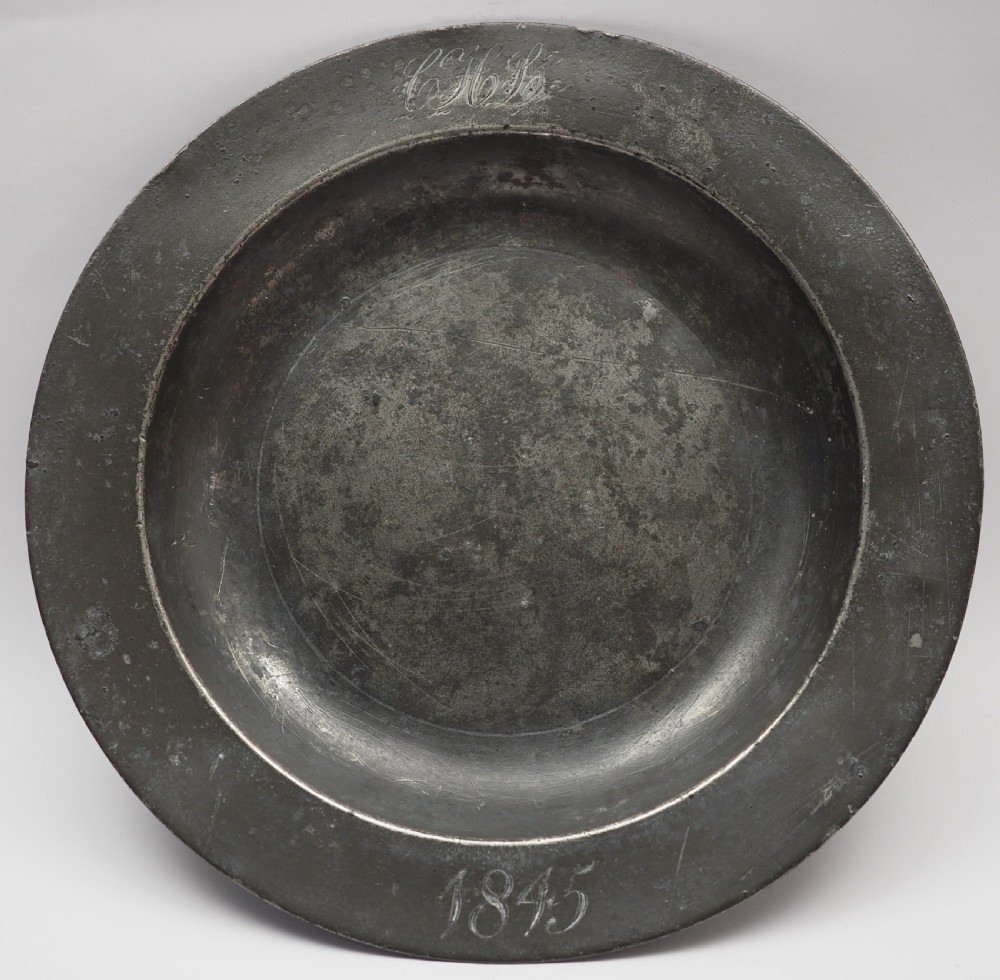 A circular grey antique pewter deep plate featuring a wide plain rim engraved with monogrammed initials at the top and the date 1845 at the bottom.