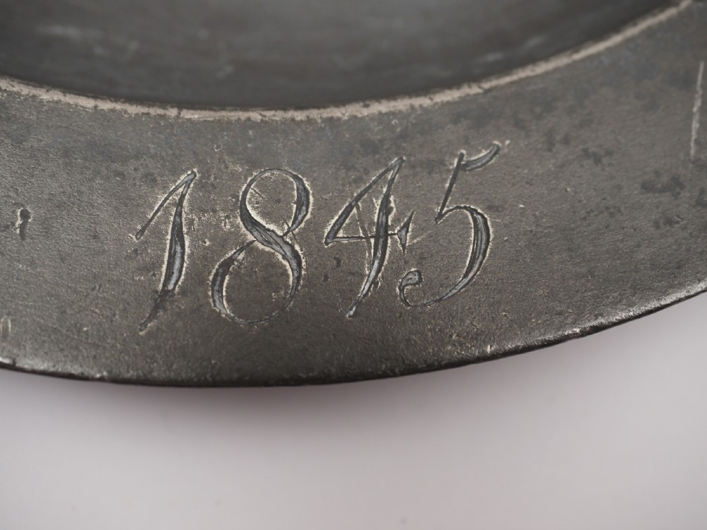 A circular grey antique pewter deep plate featuring a wide plain rim engraved with monogrammed initials at the top and the date 1845 at the bottom. View IV