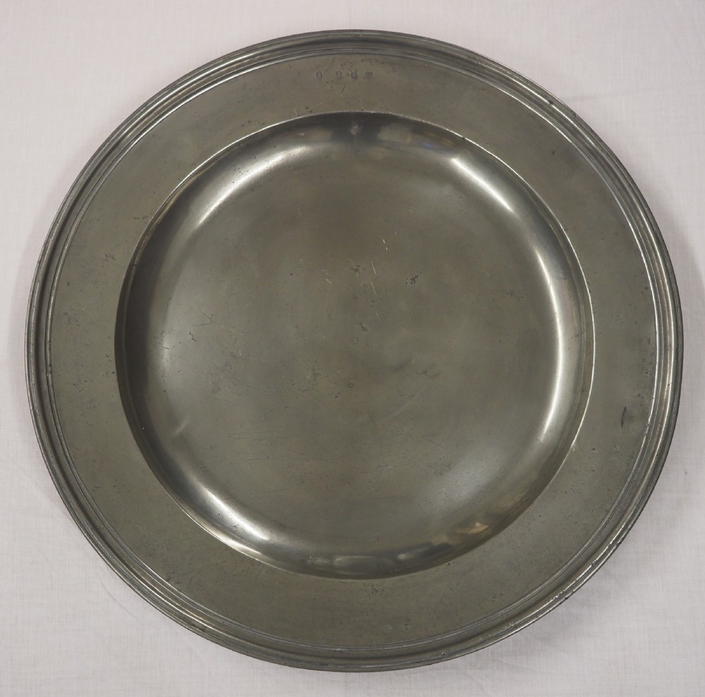 A circular antique grey pewter charger plate featuring a wide rim with reeded moulding and stamped maker touchmarks along the upper edge.