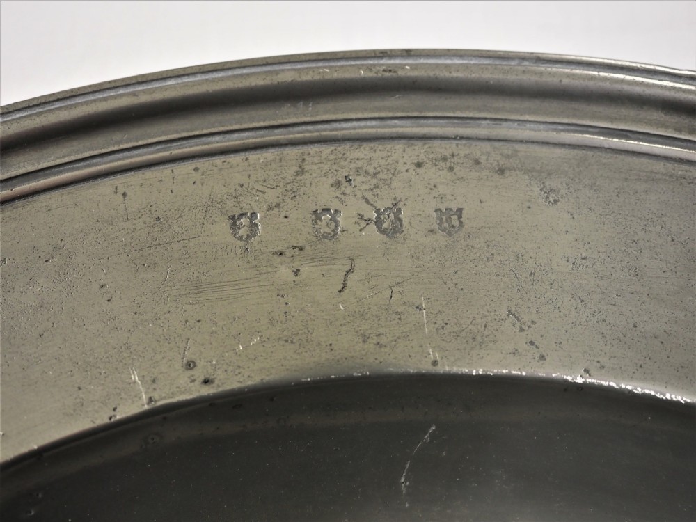 A circular antique grey pewter charger plate featuring a wide rim with reeded moulding and stamped maker touchmarks along the upper edge. View III