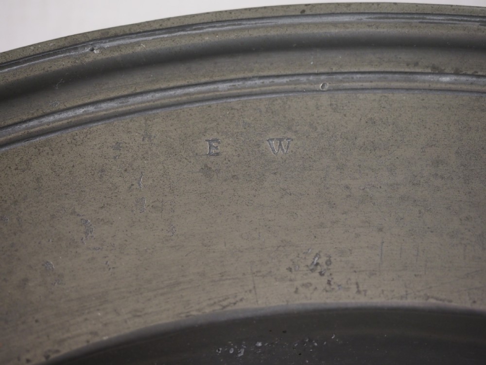 A circular antique grey pewter charger plate featuring a wide rim with reeded moulding and stamped maker touchmarks along the upper edge. View V