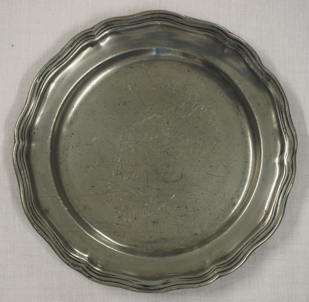 A circular antique pewter charger plate featuring a moulded wavy border and light surface knife marks.