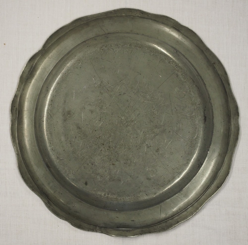 A circular antique pewter charger plate featuring a moulded wavy border and light surface knife marks. View II