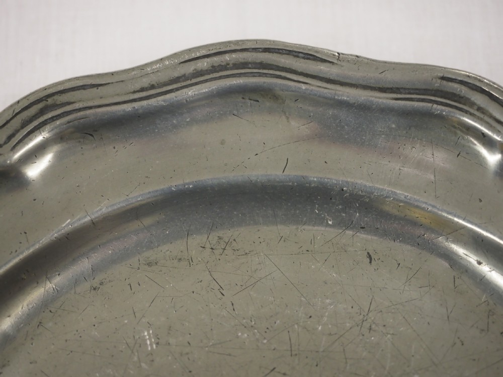 A circular antique pewter charger plate featuring a moulded wavy border and light surface knife marks. View III