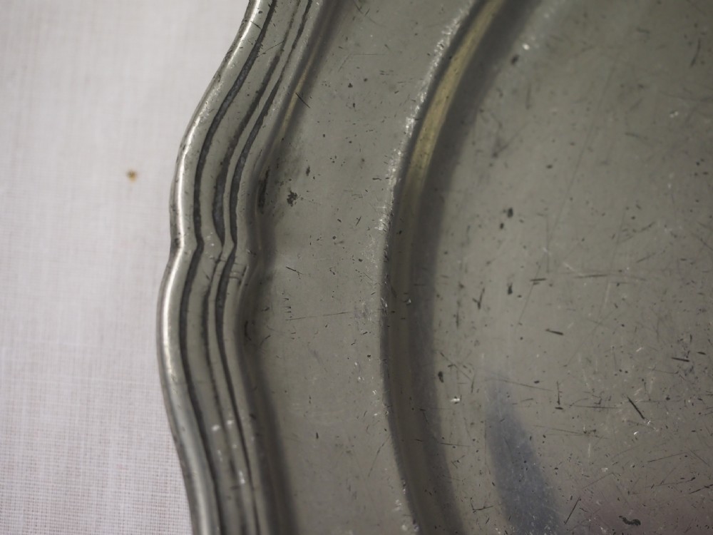 A circular antique pewter charger plate featuring a moulded wavy border and light surface knife marks. View IV